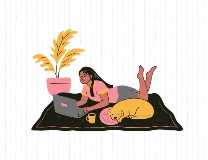 A person lies on a rug while working on a laptop and a dog sleeping beside them. A potted plant is nearby.