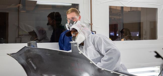 student with spray paint gun working on car bumper in paint booth
