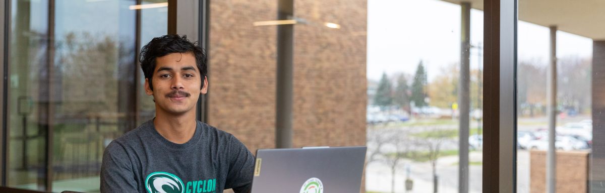 male student sits at a table with an open laptop with windows in the background at the main entrance of SCTCC