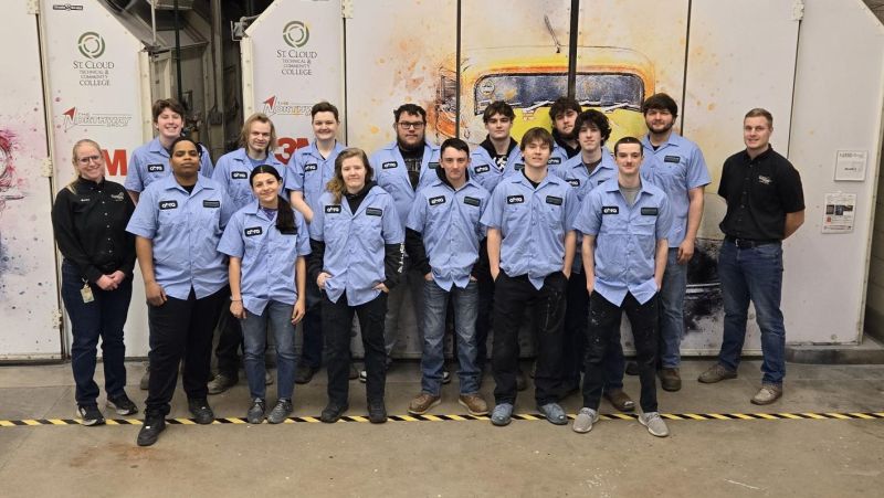 Auto body students in front of paint booths wearing donated blue shirts