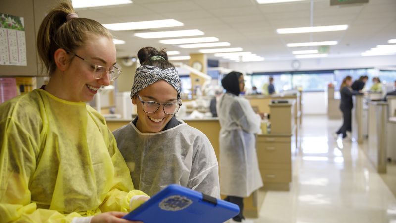 Two dental students look at a chart together with the dental clinic in the background
