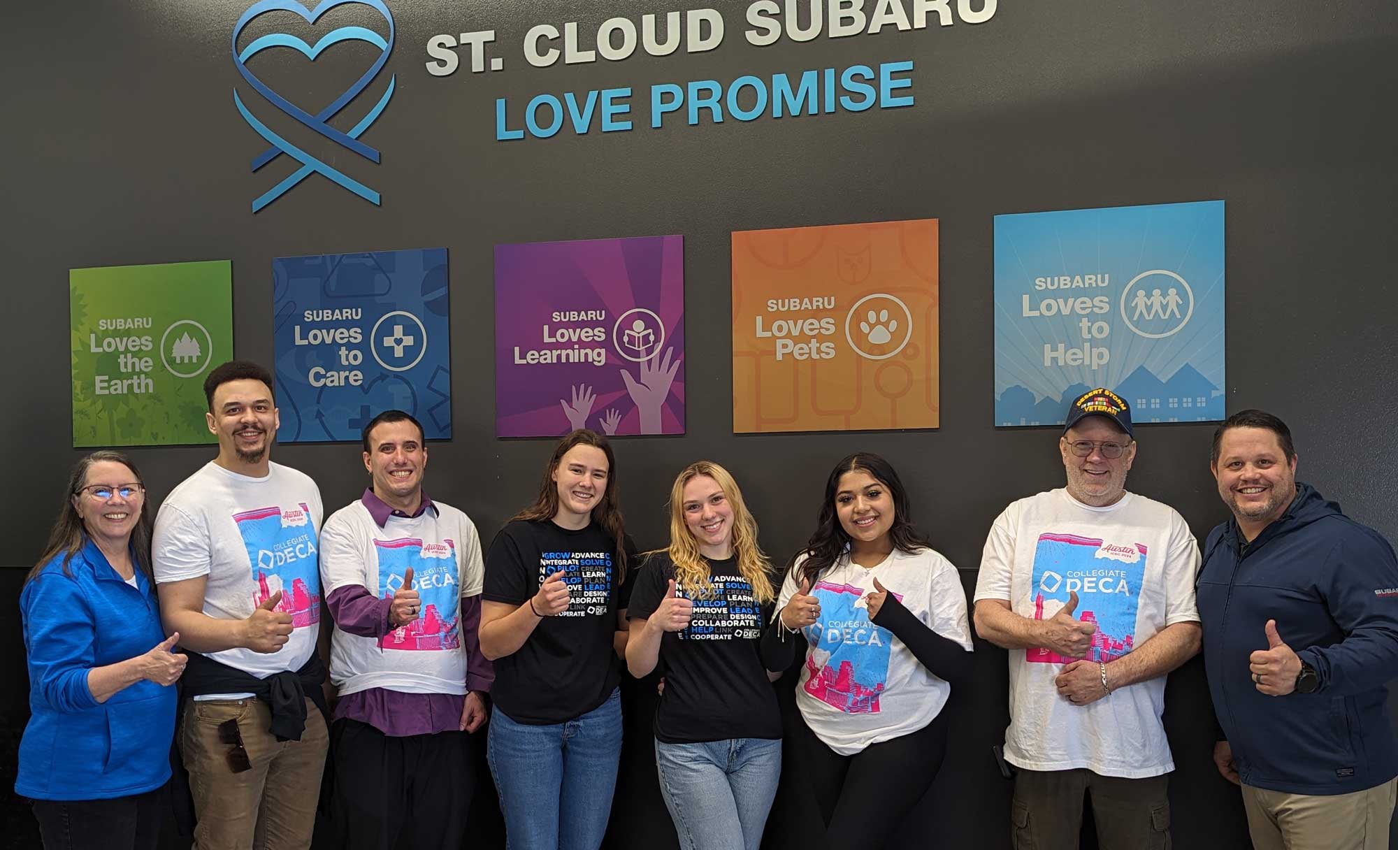 8 people in row in front of Subaru Love graphics giving thumbs up